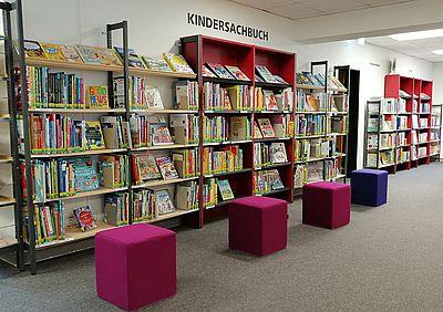 Eine lange Bücherwand mit Kinderbüchern, davor stehen 4 quadratische Stoffhocker